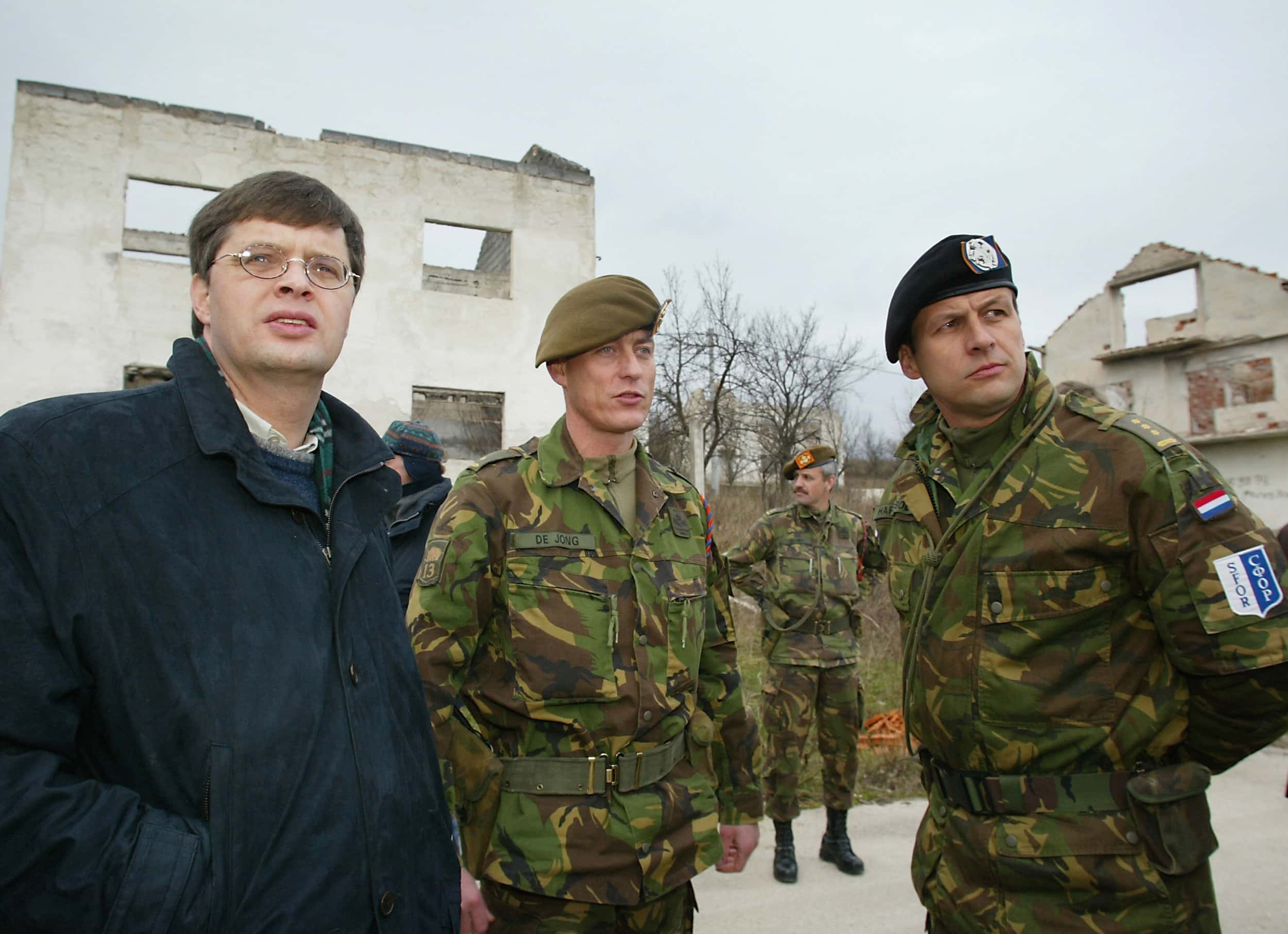 Kleurenfoto. Drie mannen zijn met elkaar in gesprek. De linker draagt een donkerblauwe jas en een bril, de andere twee zijn militairen in camouflage-uniform met baretten. Ze kijken naar iets buiten beeld. Daarachter staat een andere militair, ook in camouflage-uniform, met zijn handen op de rug. In de achtergrond ruïnes van kapotgeschoten huizen.