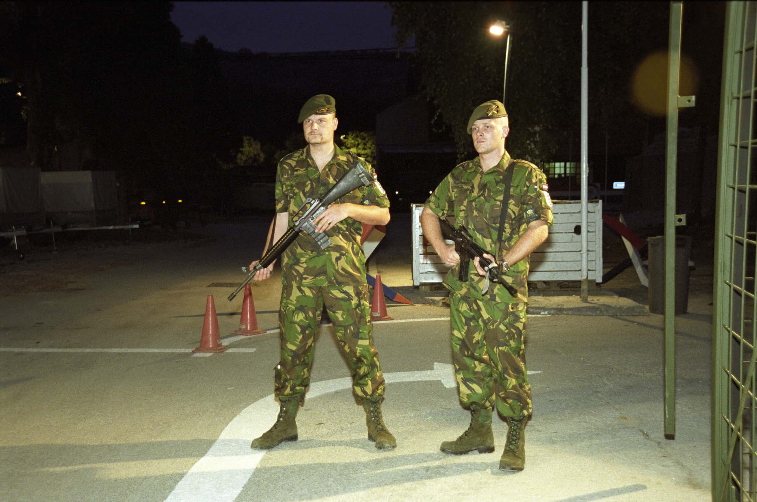 Kleurenfoto. Twee militairen staan ’s avonds op een geasfalteerde weg, een grote witte pijl naar rechts onder hun voeten, het is donker buiten. Ze dragen uniforms in camouflagekleuren, groene laarzen en een baret. Voor hun borst houden ze een geweer, ze kijken serieus naar iets links achter de camera. Helemaal rechts in de voorgrond is het einde van een groen hek te zien, achter de militairen een paar pylonen op een rijtje en een brandende lantaarnpaal. Onder de lantaarnpaal staat een grote ijzeren kist.