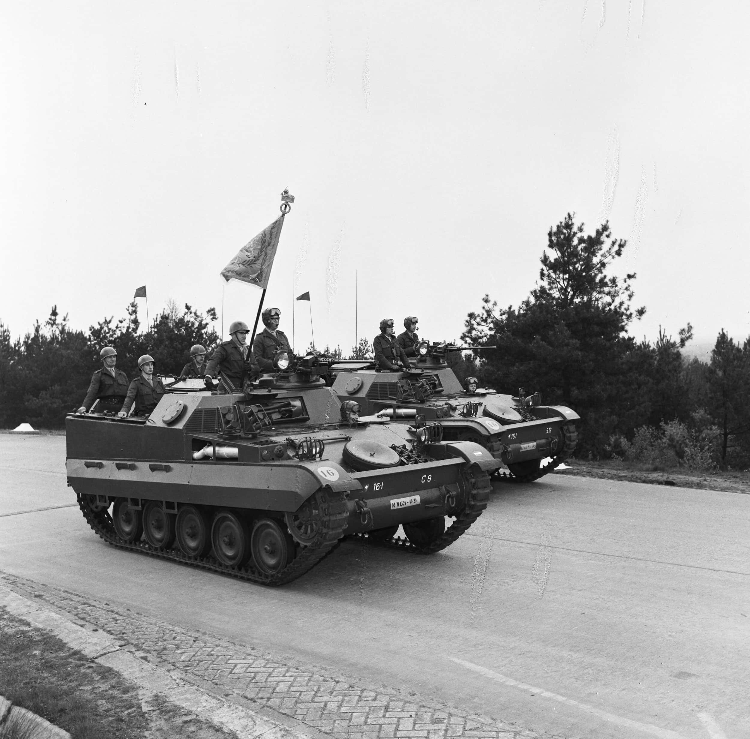 Zwart-witfoto. Twee pantserwagens rijden over een weg, uit beide wagens steken meerdere militairen uit de luiken omhoog. Ze dragen allemaal een uniforme en helmen, sommigen hebben ook een grote bril op de helm. Uit de linker wagen steekt in het middel een lange vlaggenmast omhoog, met daaraan een vierkante vlag. In de achtergrond, aan de andere kant van de weg, is een berm met bomen en struiken.