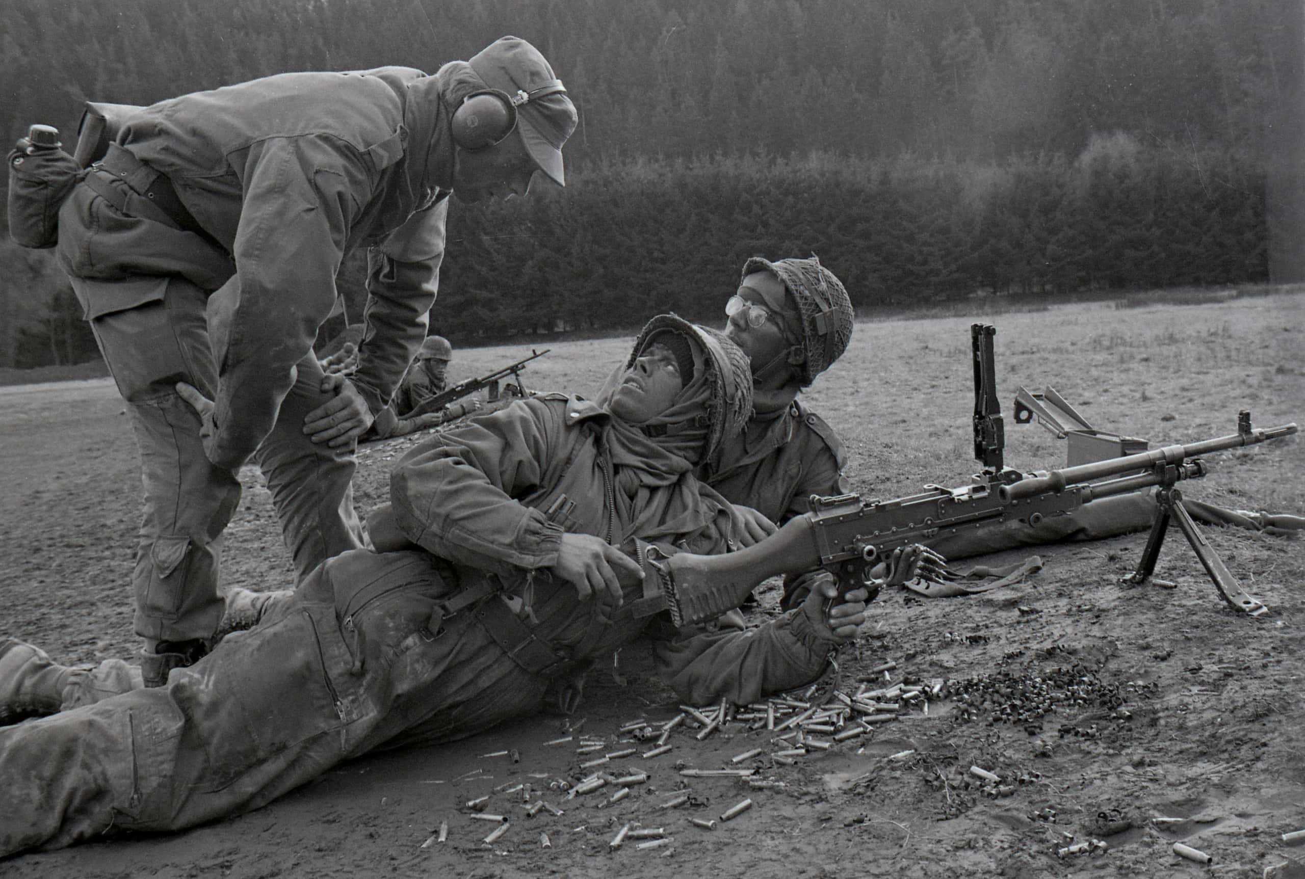 Zwart-wit foto. Er zijn drie militairen te zien, in uitrusting. Een militair staat voorovergebogen, de andere twee militairen liggen op de grond. Een van de militairen heeft een wapen.
