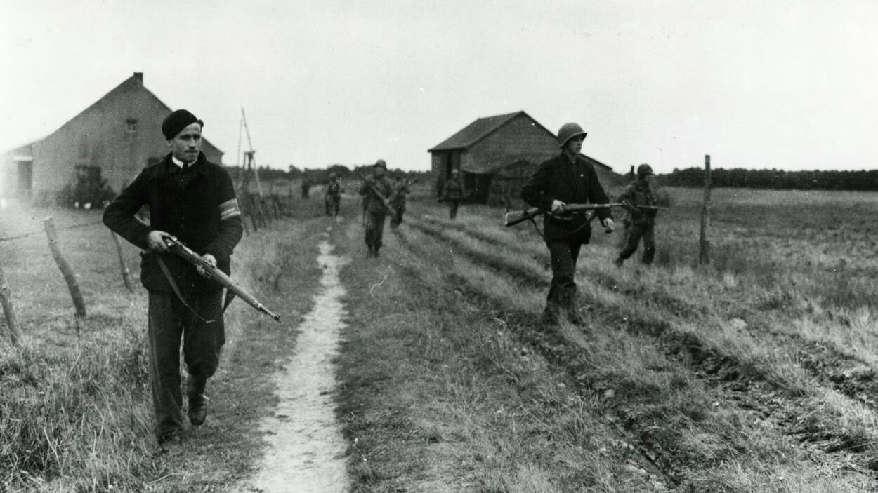 Zwart-witfoto van een Nederlandse verzetsstrijder met geweer en band om de arm, lopend over een landweg, achter hem lopen Amerikaanse militairen.