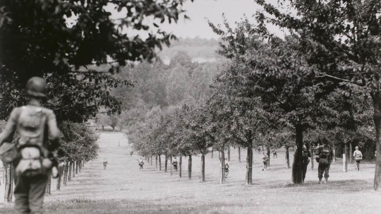 Zwart-witfoto, militairen lopen door een boomgaard richting een rivier.