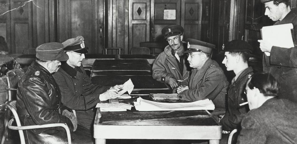 Zwart-witfoto, aan een tafel zitten links twee mannen in uniform, een van hen leest iets op van een papier in zijn handen, rechts luisteren 5 heren met documenten voor zich