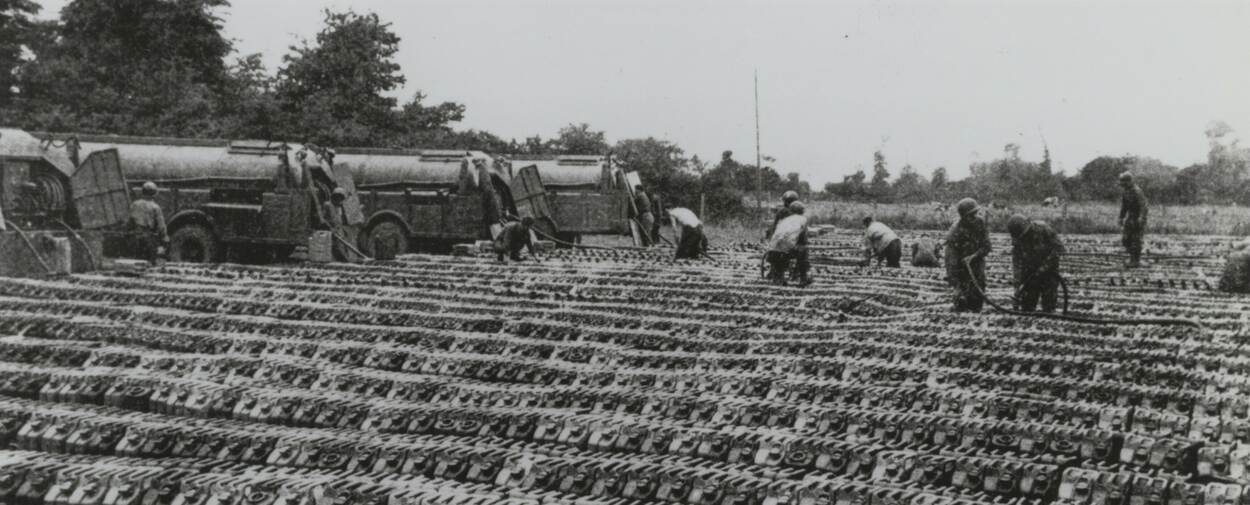 Zwart-witfoto, vele rijen jerrycans staan opgesteld, op de achtergrond vullen mannen met slangen de jerrycans vanuit 4 tankwagens.