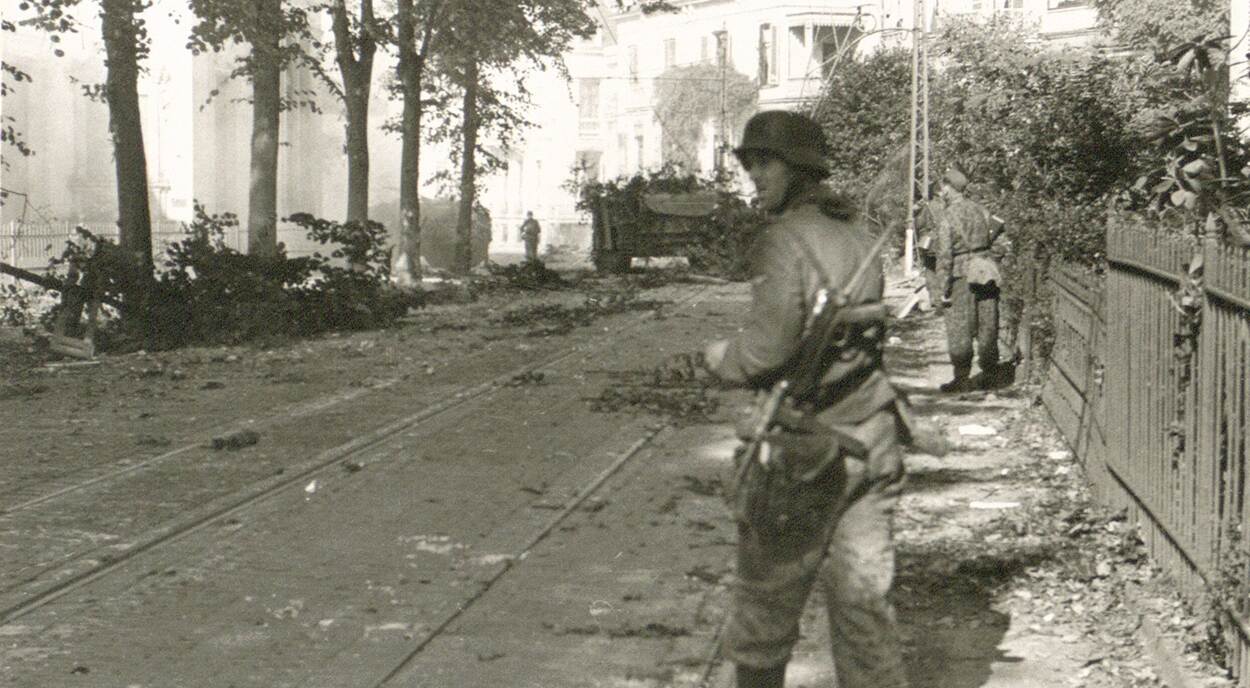 Zwart-witfoto, een straat met trambaan ligt vol puin en takken, op de voorgrond loopt een militair die achteromkijkt, op de achtergrond een tank bezaaid met takken.