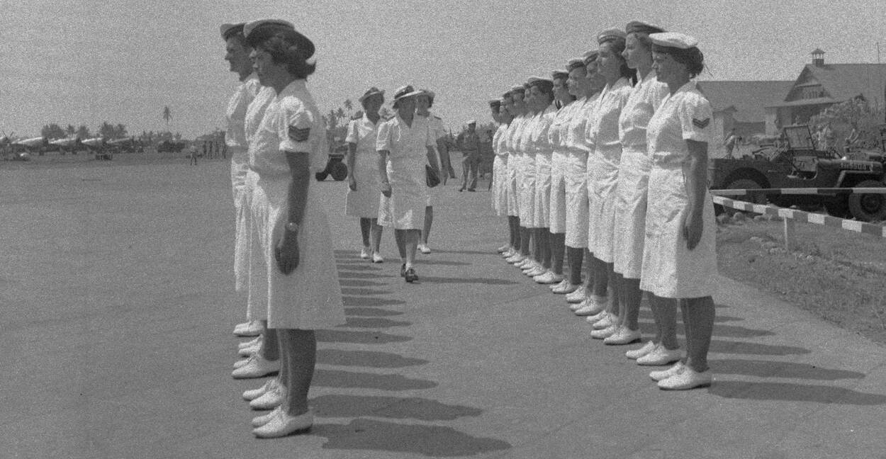 Zwart-witfoto, Van Slooten loopt samen met twee vrouwen tussen twee rijen marva's in witte uniformen op een landingsbaan. Links op de achtergrond zijn stilstaande vliegtuigen te zien.