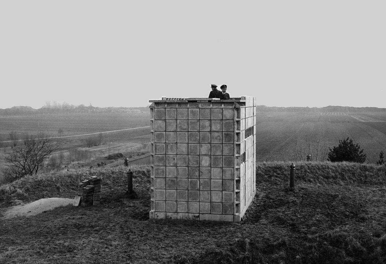 Zwart-witfoto, een kleine toren van betonnen blokken op een heuvel. Op de toren houden twee militairen de wacht.