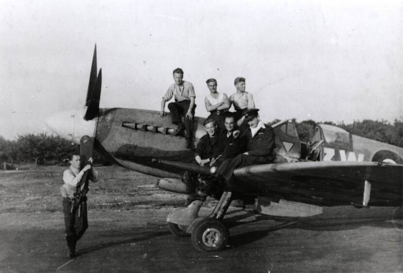 Zwart-witfoto van een propellervliegtuig, op het toestel en de vleugel zitten 6 mannen, naast de propeller staat 1 man.