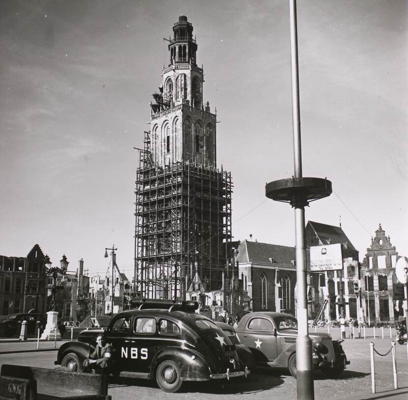 Zwart-witfoto, een auto met in witte letters 'NBS' staat op een plein geparkeerd met een kerktoren in steigers op de achtergrond. Op de zijkant van de auto zit een man met een geweer en een band om de arm.