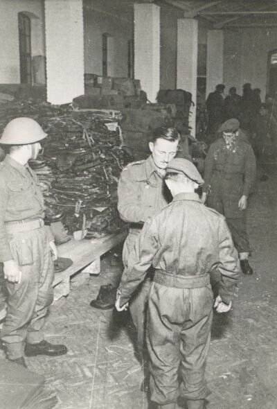 Zwart-witfoto, een Britse militair helpt een man met het dichtknopen van zijn uniform. Op de achtergrond liggen pallets met stapels kleding.