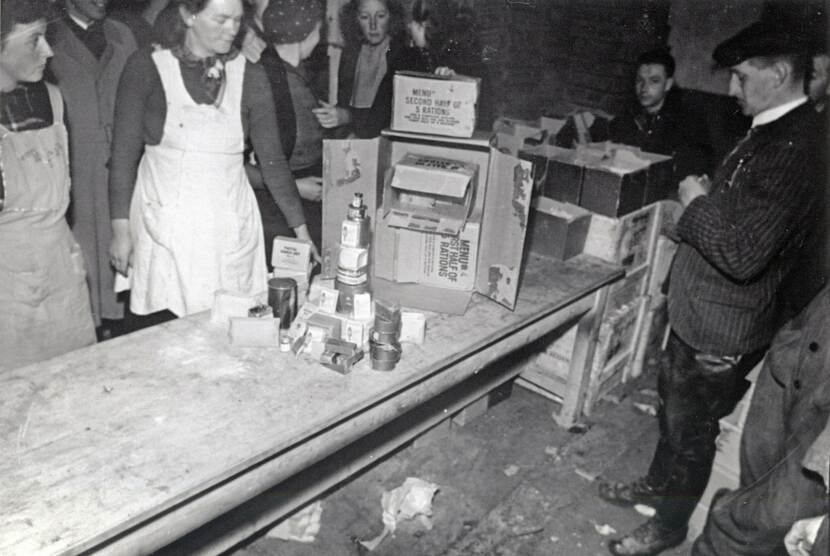 Zwart-wit foto van een ruimte met een lange houten tafel in het midden, op de tafel staan dozen met in het Engels vermeld dat het om rantsoenen gaat, en opgestapelde blikken en potten, links twee vrouwen met witte schorten,  rechts staan enkele heren in zwarte jasjes.