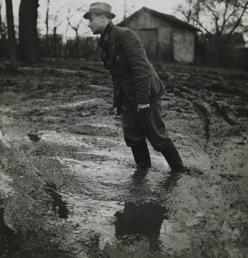 Zwart-witfoto, een man in burgerkleding staat met kaplaarzen tot zijn enkels in een modderpoel.