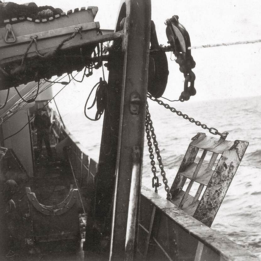 Zwart-witfoto vanaf het dek van een schip van een werktuig dat in zee wordt getakeld met een ketting.