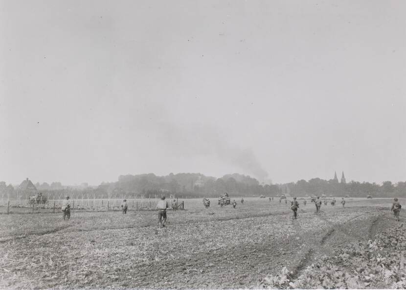 Zwart-witfoto, lopende militairen en tanks bewegen zich door een veld richting een op de achtergrond gelegen dorp.