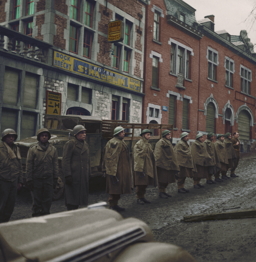 Foto genomen vanuit een auto van 12 staande militairen op een rij in een modderige straat, ze hebben helmen op en 8 mannen een regenmantel over het uniform, achter hen staat een bruine vrachtwagen met open laadruimte geparkeerd, op de voorgrond de neus van de donkergroene auto.