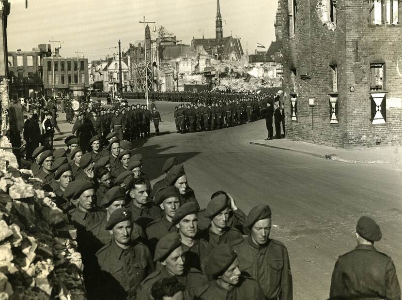 Zwart-witfoto, tientallen militairen marcheren door verwoest Groningen.