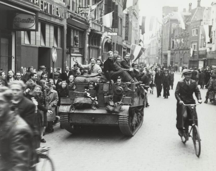 Zwart-witfoto, jonge mensen op een brencarrier lachen en zwaaien naar de camera in een drukke straat.