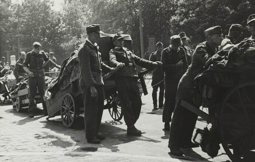 Zwart-witfoto van een stoet militairen met volgeladen karren in een straat, de karren hebben 2 wielen en worden met de hand getrokken aan de voorkant, en geduwd aan de achterkant.