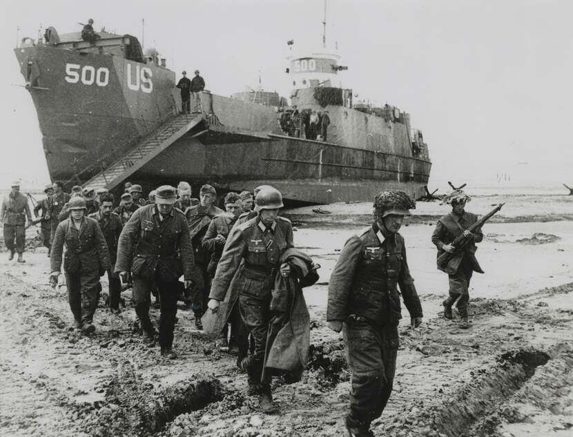 Zwart-witfoto, een groep militairen loopt weg van een schip met opschrift '500 US', op het schip kijken mannen toe vanaf het dek, rechts escorteert een militair met een geweer de groep.