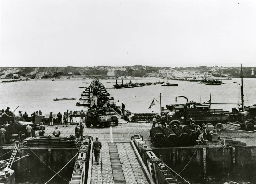 Zwart-witfoto, een pierhoofd op zee met voertuigen en militairen, vanaf het pierhoofd loopt een drijvende brug op pontons richting het strand, hier overheen rijden voertuigen.