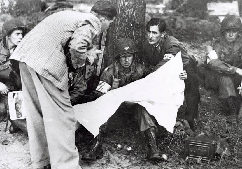 Zwart-witfoto, een militair met helm zit tegen een boom een kaart te lezen, naast hem staan twee mannen in burgerkleding hem te helpen.