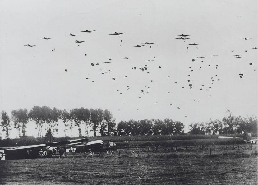 Zwart-witfoto, in de lucht veel vliegtuigen en parachutisten, onderin een veld met 2 vliegtuigen.