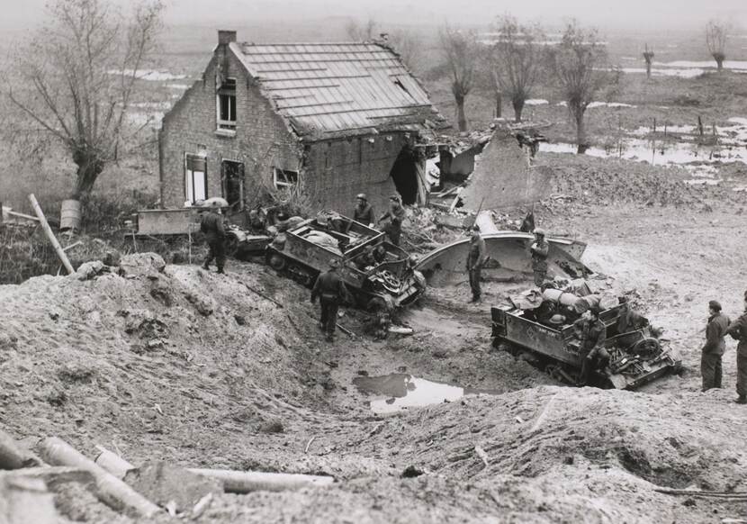 Zwart-witfoto van pantserwagens die langs een verwoest huis in een weiland rijden.