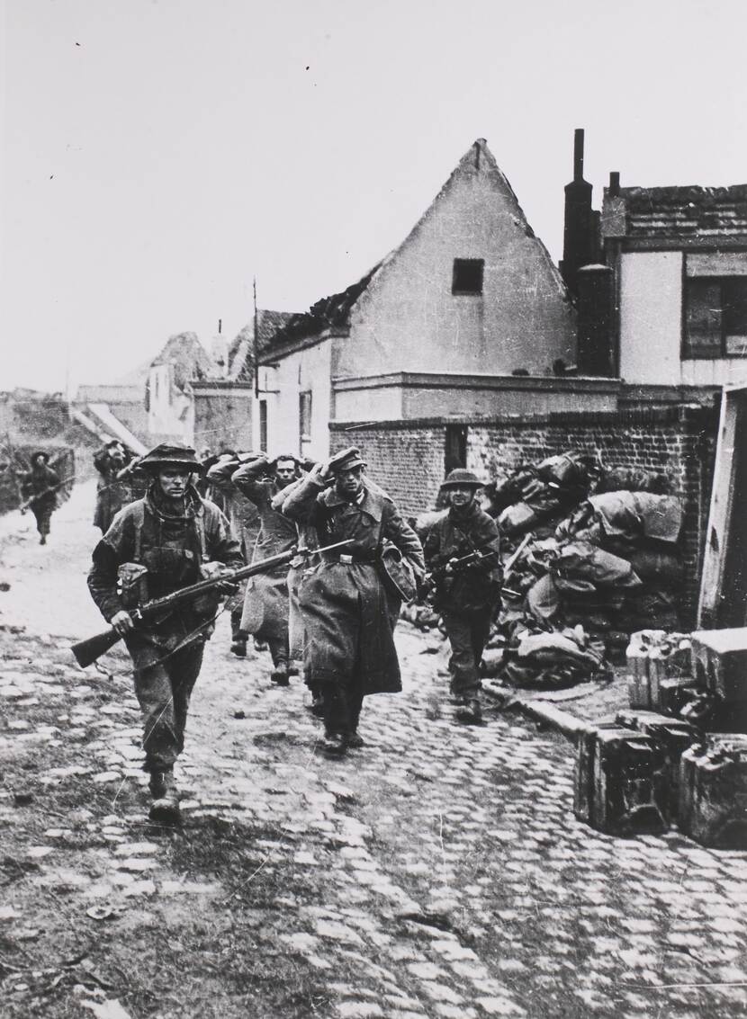 Zwart-witfoto, Duitse militairen lopen in een rij, met de handen op het hoofd, door een verwoeste straat, begeleid door gewapende Franse militairen.