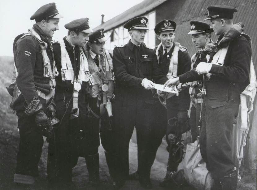 Zwart-wit foto met 7 militairen in uniform. Ze zijn in overleg en bekijken een notitie.