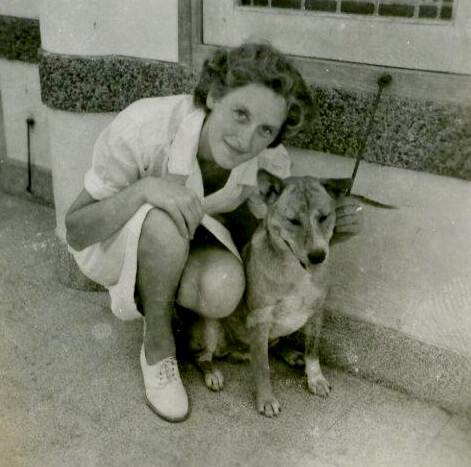 Zwart-witfoto van een jonge vrouw in wit marine-uniform knielend naast een hond.