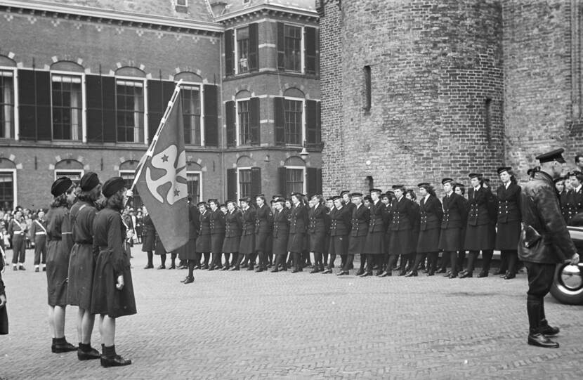 Zwart-witfoto, op de voorgrond staan 3 padvindsters met een vaandel. Daartegenover staat een erewacht van marva's aangetreden in donker uniform.