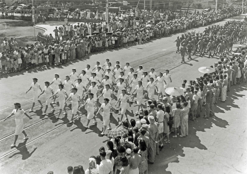 Zwart-witfoto van een militaire parade. Een detachement marva's marcheert in witte uniformen met langs de weg veel toeschouwers.