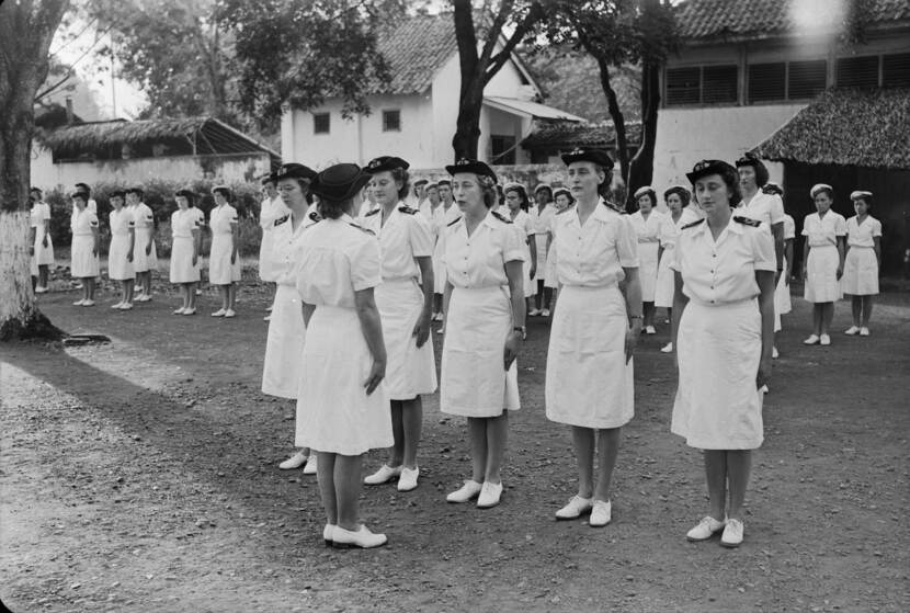 Zwart-witfoto, op een binnenplaats staan marva's aangetreden in wit uniform. 5 vrouwen staan op de voorgrond met daarvoor Wibbine Verschuren op de rug gezien.