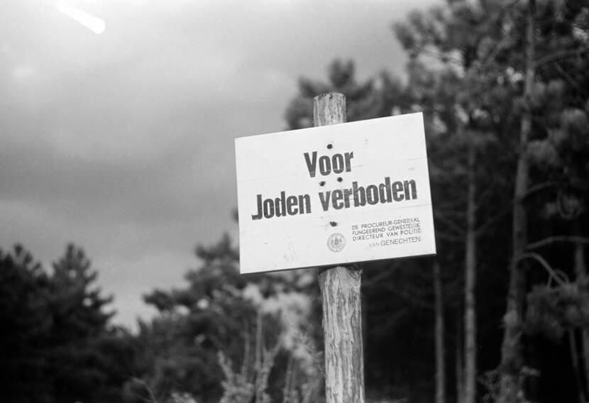 Zwart-wit foto. Bord met de tekst ‘Voor Joden verboden’. Het witte bord is bevestigd op een paal, op de achtergrond zijn bomen te zien.