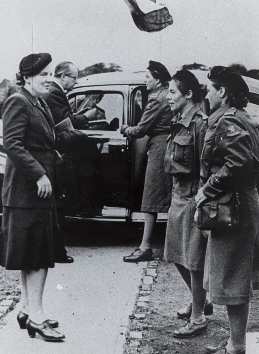 Zwart-wit foto. Koningin Juliana wordt ontvangen door twee vrouwelijke militairen. Op de achtergrond is een auto te zien.