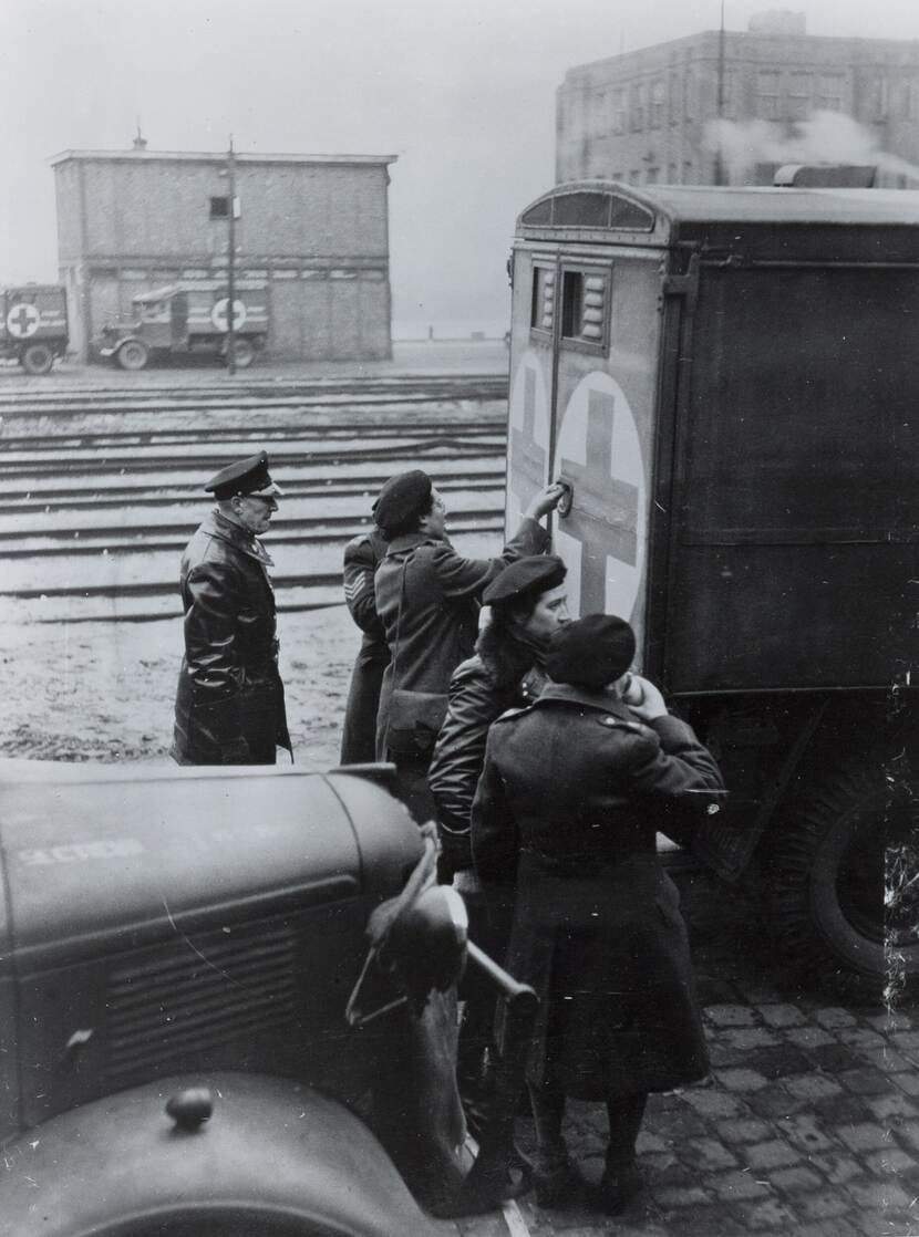 Zwart-wit foto. Een aantal militairen staan bij de achterzijde van een ambulance. Op de achtergrond ligt treinrails en staan nog meer militaire voertuigen.