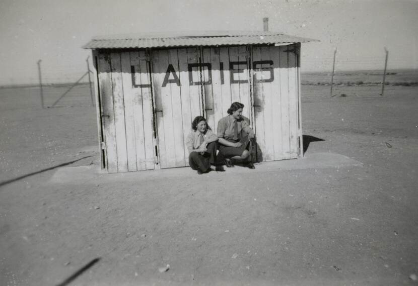 Zwart-witfoto van twee vrouwen in uniform voor een houten huisje met 'Ladies' erop geschilderd. Ze bevinden zich in een woestijnachtige omgeving.