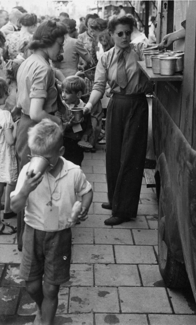 Zwart-witfoto, twee vrouwen in militair uniform met broek en stropdas delen vanaf een voedseltruck bekers uit aan kinderen.