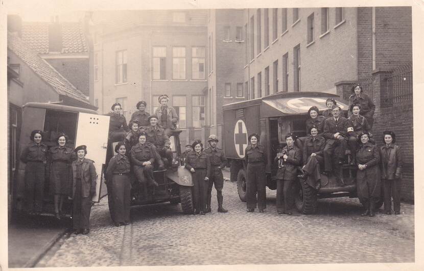 Zwart-witfoto, 2 ambulances en 1 jeep staan in een straat met ervoor/erop leden van het VHK.