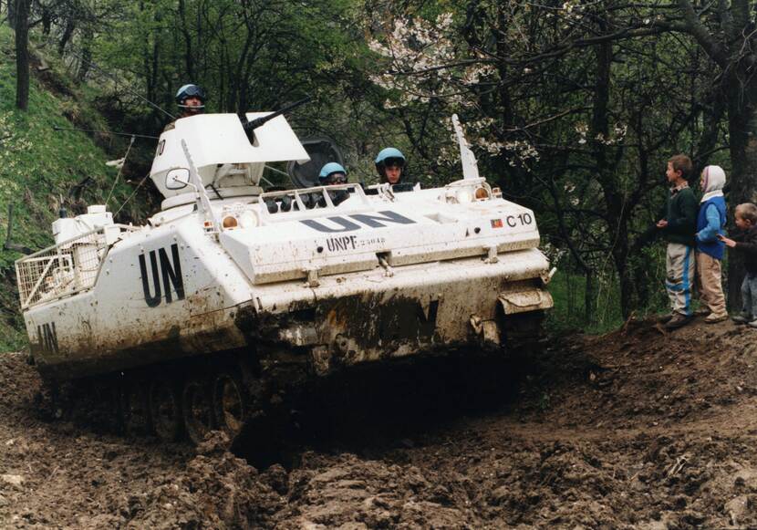 Een witte pantserwagen rijdt door de modder in een bos, in de wagen zitten 3 militairen met blauwe helmen op, rechts staan 3 jonge kinderen toe te kijken.