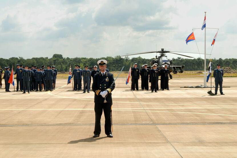 Militairen staan opgesteld in ceremonieel tenue op de paradeplaats, op de achtergrond staat een helikopter.