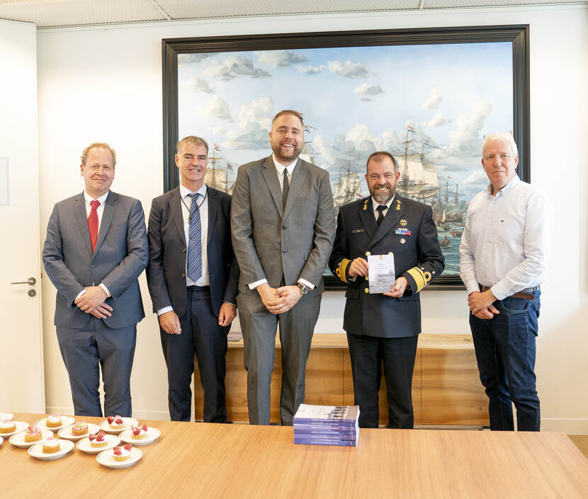 Groep poseert voor voor een schilderij met varende schepen. Drie mannen in pak, een man in militair unform en een man in een wit overhemd. Voor hen liggen boeken.