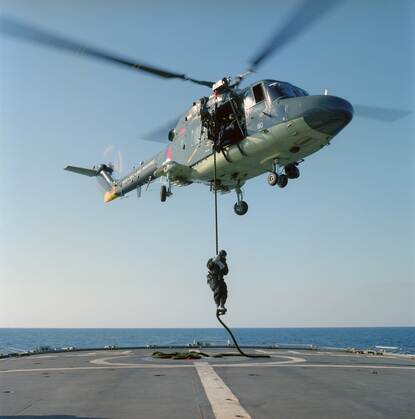 Foto vanaf het helikopterdek, een helikopter zweeft een stuk boven het dek, uit de helikopter hangt een kabel waarin een militair hangt die stevig vasthoudt, op de achtergrond is zee te zien.